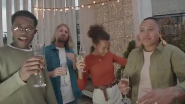 Elinde şampanya flütleri olan neşeli çoklu etnik gençlerin el kamerasıyla terasta coşkulu bir şekilde dans ederken çekilen el kamerası görüntüleri.
