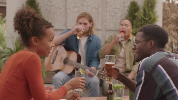 夏日的傍晚 四个无忧无虑的朋友坐在天台上 一边喝着鸡尾酒 一边喝着啤酒 会弹奏吉他的年轻人 — 图库视频影像