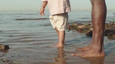 Sahilde çıplak ayakla yürüyen ve Afro-Amerikan bir babayı elle tutan tanınmayan bir çocuğun düşük kesimli yavaş çekimi.