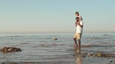 Yavaş çekimde Afrikalı-Amerikalı bir adam kucağında yeni yürümeye başlayan oğlunu omuzlarında taşıyarak güzel sahil şeridinde yürüyordu.