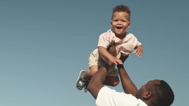 一个深情的年轻非洲裔美国男子在户外海滩度过阳光灿烂的一天 从低角度慢镜头向空中抛掷快乐的幼儿 — 图库视频影像