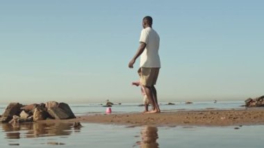 Gün ışığı altında güzel kumsalda el ele dönen, sevimli bir çocuğu tutan, gündelik giysili, mutlu Afro-Amerikan bir adamla geniş açılı çekim.