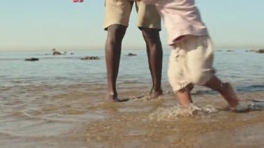 Afrika kökenli Amerikalı baba ve oğlunun yaz günü kıyı şeridi boyunca suda koşarken çekilmiş yavaş çekimleri.