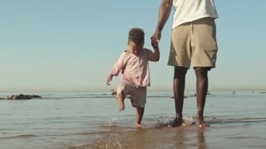 Şirin Afro-Amerikalı çocuk babasıyla birlikte güzel deniz kıyısında yürüyor, ayağıyla berrak mavi su sıçrıyor ve birlikte eğleniyorlar.