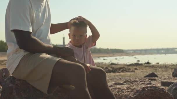 若いですショットのアフリカ系アメリカ人男Patting彼のかわいいです2歳の息子で頭に一緒に座って砂浜で日光浴 — ストック動画