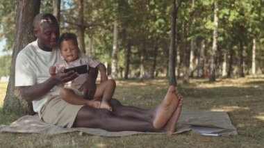 Sevecen Afro-Amerikan babasının tatlı çocuğuna komik videolar gösterirken yavaş çekimde güneşli bir günde, ağaçların altında battaniyenin altında otururken.