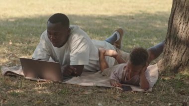 Afro-Amerikalı bir adam parkta, ağaçların altında yatarken dizüstü bilgisayarında video konferansı yapıyor. Bu sırada küçük oğlu da yakınlarda uzanmış akıllı telefondan videolar izliyor ve birlikte yaz tatilinin tadını çıkarıyor.
