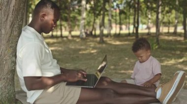 2 yaşındaki oğlu yakınlarda oynarken, ağaçların altında dizüstü bilgisayarla çalışan Afro-Amerikan bir adamın yan görüntüsü.
