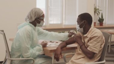 Koruyucu maske takmış, giysi giymiş, genç Afrikalı Amerikalı bir adamın kolunda Covid-19 aşısı yaparken orta boy bir fotoğraf.