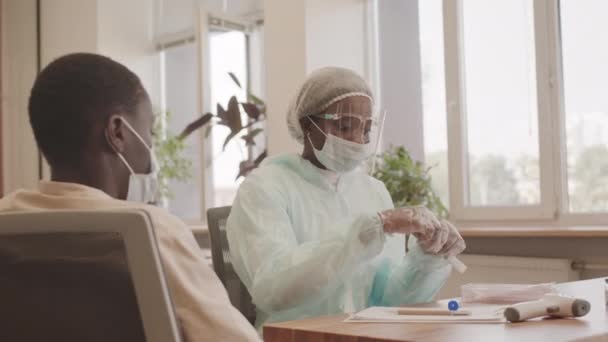 ミディアムショットの女性医療従事者で保護服や顔シールド収集喉スワブからアフリカ系アメリカ人男性取得テストのためにCovid — ストック動画