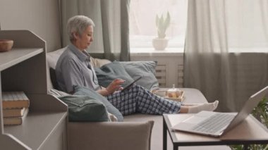 Beyaz saçlı, pijama giyen, evdeki kanepede oturup tablet bilgisayar kullanan Asyalı kadının yan görüntüsü.