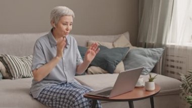 Orta uzunlukta, pijama giyen, kablosuz kulaklık takan, oturma odasında koltukta oturan, taşınabilir bilgisayar kullanan Asyalı bir kadın.