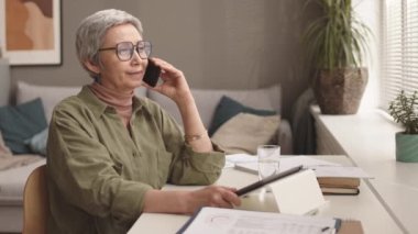 Modern, üst düzey Asyalı bir kadının rahat ofisinde masasında oturup tablet bilgisayarla çalışırken cep telefonuyla konuşurken belini kaldırması.