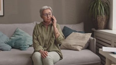 Orta uzunlukta başarılı bir Asyalı kadın ev ofisindeki koltukta oturuyor, cep telefonuyla konuşuyor, sonra ayağa kalkıyor ve masa masa değiştirip belgelere bakıyor.