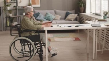 Beyaz saçlı Asyalı kadının oturma odasında masasında oturmuş tablet bilgisayar kullanarak çalışmasının tam kenar görüntüsü.