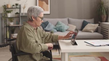 Oturma odasında tekerlekli sandalyede oturan, tablet bilgisayar kullanarak çalışan yaşlı Asyalı kadının orta boy yan görüşü.