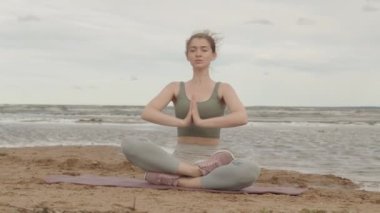 Sabahları yogayı tek başına yapan beyaz bir kadının yavaş çekimleri. Nilüfer pozisyonunda oturmuş, kumlu sahilde yoga minderinde dua eden ellerle su kenarında.