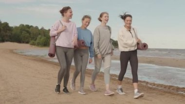 Spor kıyafetli, yoga minderleriyle kumsalda yürüyen ve sohbet eden dört zayıf kadının yavaş çekimleri.