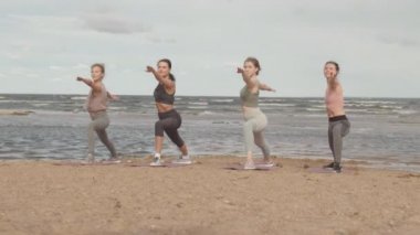 Spor giyimli dört beyaz kadının kumlu sahilde sırayla durup yoga yaparken, savaşçı 2 pozunda durduğu uzun soluklu, yavaş çekim.