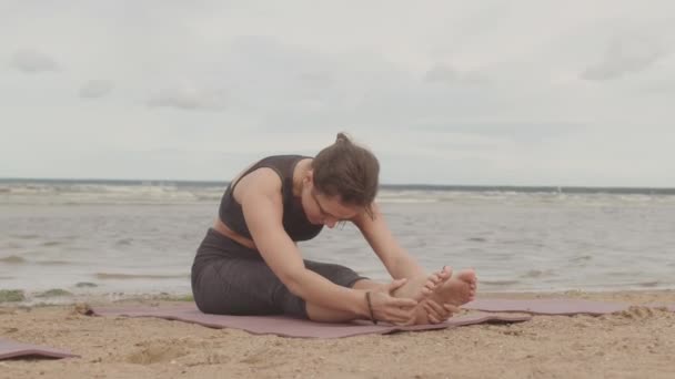 スポーツウェアで若いリラックスした女性のスローショット目を閉じて砂浜に一人で座ってストレッチ演習を行う リラックスして自然を楽しむ — ストック動画