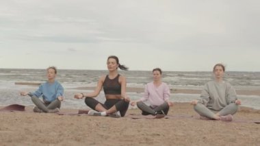 Bulutlu ve rüzgarlı bir günde, sıcak spor kıyafetleriyle kumlu sahil şeridinde yoga yapan dört genç kadının, Lotus pozunda yoga minderleri üzerinde meditasyon yapmasının yavaş çekimi.