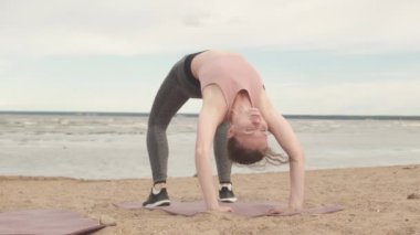 Konsantre olmuş genç bir kadının briç pozisyonunda rüzgarlı ve bulutlu bir günde ıssız bir sahilde yoga yaptığı yavaş çekim.