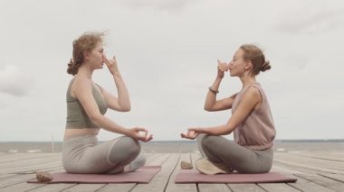 İki genç kadının Lotus 'ta oturup birbirlerinin önündeki ahşap iskelede poz verirken, meditasyon yaparken ve alternatif burun deliği nefes alma tekniği uygularken yavaş çekim.