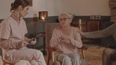 Yaşlı bir kadının artritten muzdarip olduğu yavaş çekim. Genç destekçi hemşireyle konuşuyor. Panoya not alıyor.