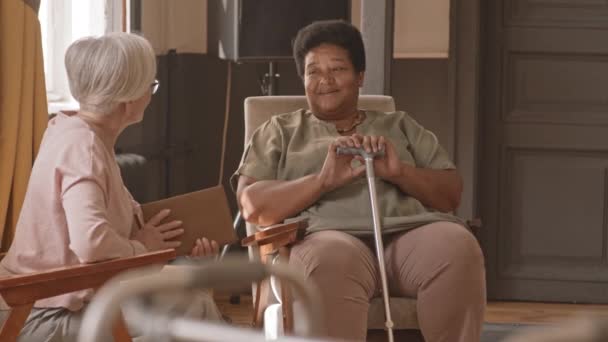 介護施設で会話をしている白人とアフリカ系アメリカ人の高齢者の女性がゆっくりと撮影された 看護師は彼らの世話をし立ち上がって歩くのを助け — ストック動画