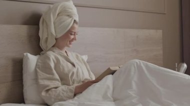 Kafasında havluyla bornoz giymiş, beyaz yatakta oturmuş romantik kitaplar okuyan hayalperest bir kadın resmi.