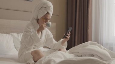 Beyaz bornozlu, kafasında havluyla, altın rengi göz bantlarıyla, sabah kahvesinin tadını çıkarırken ve akıllı telefonuyla yatakta otururken orta boy beyaz bir kadın resmi.