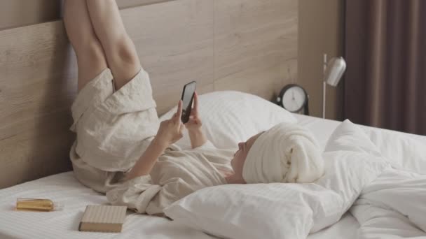 Mulher Branca Jovem Roupão Banho Toalha Cabeça Rolando Smartphone Deitado — Vídeo de Stock