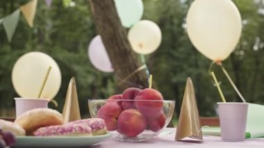 Şeftalili kase, bir tabak donut, pipetli kağıt bardaklar ve açık havada parti şapkaları, rüzgarda savrulan hava balonları.