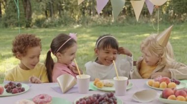Beş yaşında çeşitli çocuklar doğum günü partisi veriyor, gıdıklanıyor, gülümsüyor ve güneşli bahçede masada oturuyor.