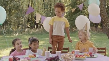 Orta boylu kıvırcık saçlı, sandalyeye oturmuş, arkadaşları yaz parkındaki doğum günü partisinde masada oturuyor.