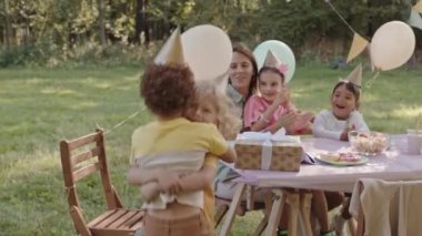 Sarışın, mutlu bir çocuk. Karışık ırk arkadaşını kucaklıyor ve kaldırıyor. Parti şapkaları takıyor, kadınlar ve kızlar yaz parkındaki doğum günü partisinde masada oturuyor.