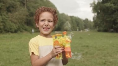 Orta boylu kıvırcık ırklı erkek ilkokul öğrencisi. Sunny Park 'ta oyuncak sabun yapımcısıyla oynarken gülümsüyor.