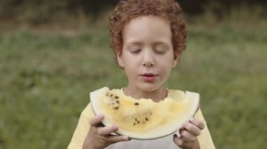 Açık havada karpuz yiyen, kameraya bakan kıvırcık, şirin, kıvırcık bir çocuğun göğüs portresi.