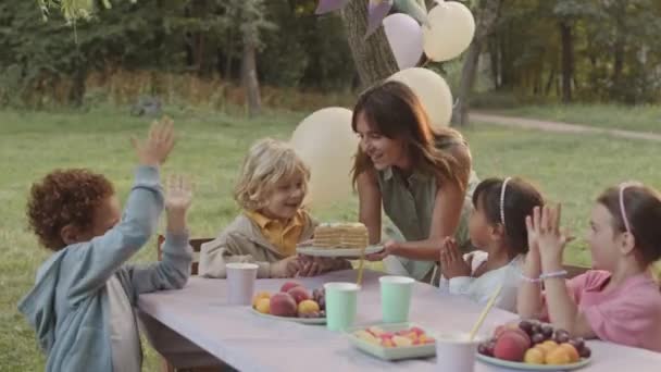 中長いです若いです白人の母親サービング誕生日ケーキへ彼女の5歳ブロンド息子接吻彼に 子供Clapping手座っているテーブルの上に公園 — ストック動画