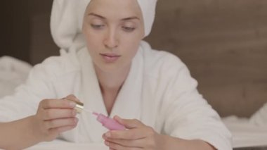 Kafasına pembe dudak parlatıcısı süren bornoz ve havlu takan genç bir kadının orta boy yakın çekimini yapıp rahat yatağında uzanan kameraya bakarak.