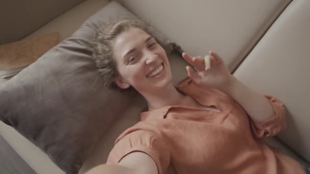 自らを記録しながらカメラに手を振る若い陽気な女性の手を振ったり 自宅でソファの上に横になってビデオチャットをしたり — ストック動画