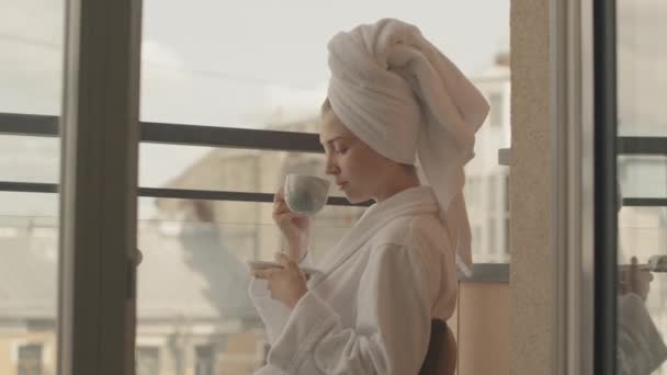 身穿白色浴衣 头戴毛巾 站在全景窗前 端着一杯咖啡的漂亮白人年轻女子的蜡像 然后转身对着镜头微笑 — 图库视频影像