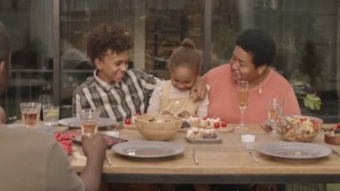 Yaşlı Afrikalı kadın şenlikli yemek masasında oturuyor, gülümsüyor ve beş yaşındaki tatlı torunu ve genç torunuyla konuşuyor.