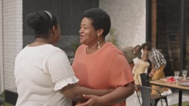 Orta yaşlı Afrikalı bir kadın olgun annesine sarılıyor, gülümsüyor, konuşuyor ve kameraya bakıyor, diğer aile üyeleri terasın arka planında duruyorlar.