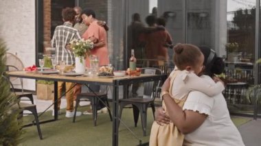 Orta uzunlukta Afrikalı bir kadın terasta 5 yaşındaki tatlı kızına sarılıyor aile üyeleri arka planda iletişim kuruyor.