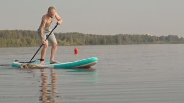 Yavaş çekimde, kolunda dövmesi olan genç bir adamın gölde kürek çekişini izliyorum.