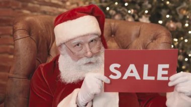 Noel Baba 'nın koltukta oturup üzerinde satılık yazı olan kırmızı tabelayı kaldırması ve kameraya bakıp gülümsemesi.