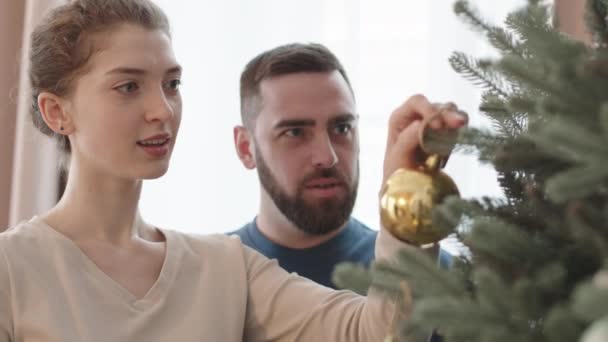 家で木の上に黄金のクリスマスボールを置く若い白人女性のクローズアップ 彼女の夫は見て笑顔 — ストック動画