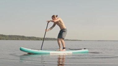 Yaz günü gölde tek başına kürek çekerken sup kürek tahtasından düşen adamın yavaş çekimleri.