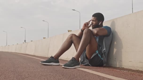 年轻的非洲裔美国运动员坐在跑道上 在慢跑前休息 参加室外晨练 在无线耳机中欣赏音乐的慢镜头 — 图库视频影像
